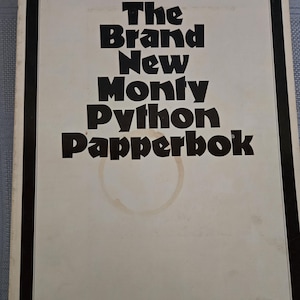 Op de afbeelding: Een vintage boekomslag met de titel "The Brand New Monty Python Papperbok" in vette zwarte letters. De omslag is gebroken wit met een zwarte rand en een cirkelvormige vlek.