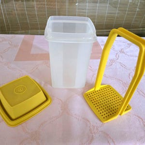 May include: A clear plastic container with a lid, a yellow square container with a lid, and a yellow potato masher. The items are on a pink surface.