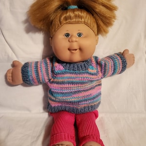 May include: A Cabbage Patch Kid doll with blonde hair in a ponytail, wearing a blue and pink striped sweater and pink trousers.