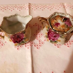 Könnte beinhalten: Ein antikes Porzellan-Sahne- und Zuckerset mit Blumenmuster. Das Set zeigt rosa Rosen und goldene Akzente.
