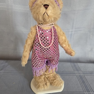 Peut inclure: Un ours en peluche vintage vêtu d'un short à paillettes roses avec des franges violettes et un collier de perles. L'ours a un bandeau violet et se tient sur une base blanche. La fourrure de l'ours est brun clair.