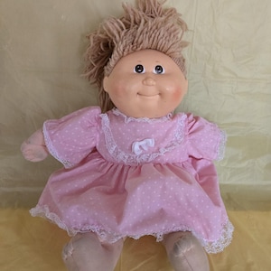 May include: A vintage doll with a round face, rosy cheeks, and brown yarn hair. The doll wears a pink dress with white polka dots and lace trim. The doll's feet are a light tan colour.