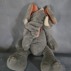 May include: A plush elephant toy featuring soft gray fur, large ears with peach-colored inner lining, and white tusks. The elephant has a long, textured trunk, dark eyes, and a small floral-patterned fabric bow attached between the ears.
