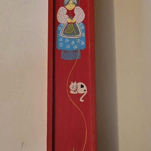 May include: A red wooden box with a painted scene of a woman knitting in a chair with a cat and a ball of yarn. The woman is wearing a blue dress with white flowers and a white hat. The cat is white with black spots. The ball of yarn is yellow.
