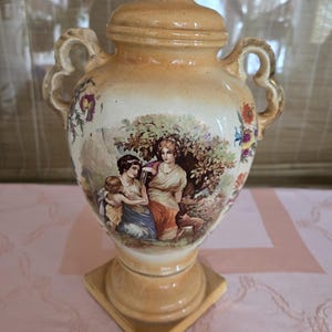 May include: A ceramic vase with a lid, featuring a painted scene of three women in a garden setting. The vase has two handles and a square base. The vase is decorated with floral motifs and is in a cream and gold colour.