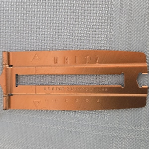 May include: A copper-coloured metal sewing gauge with a hinged design. The tool features the embossed text "DRITZ" and "U.S.A. PAT. 2939186 OR 2939187" along with a central rectangular slot and small triangular markings for precise measurement.