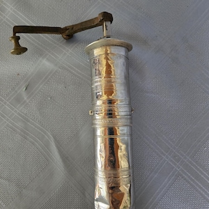 May include: A cylindrical silver-coloured metal pepper mill featuring a long, dark-toned metal hand crank at the top. The body of the mill is decorated with horizontal engraved bands and has a reflective, slightly dented surface texture throughout.