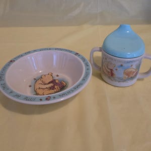 May include: A Winnie the Pooh themed baby bowl and sippy cup set. The bowl has a blue rim with text and a cartoon image of Pooh. The sippy cup has a blue lid and two handles, with a Pooh illustration.