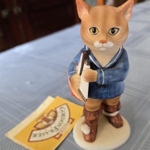 Puede incluir: Una figurita de cerámica de un gato vestido de marinero sosteniendo un salvavidas rojo y blanco a rayas. La figurita está de pie sobre una base blanca y tiene una etiqueta amarilla con el texto "Gordon Fraser Collector's Edition".