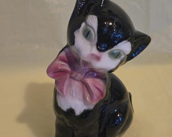 May include: A glossy black and white ceramic cat figurine with a pink bow. The cat has green eyes and is in a seated position, with its head tilted. The figurine is a decorative item.