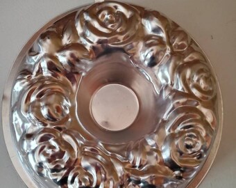 Puede incluir: Molde para pastel de metal plateado con diseño de rosas. El molde tiene forma de anillo con un agujero en el centro y un lazo para colgar en la parte superior.