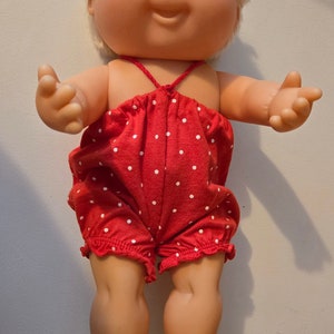 Könnte beinhalten: Eine Cabbage Patch Kid Puppe mit blonden Haaren und blauen Augen, die ein rot-weiß gepunktetes Einteiler trägt.