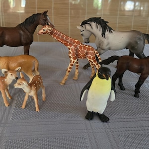 Può includere: Una collezione di figurine di animali giocattolo, tra cui cavalli, cervi, una giraffa e un pinguino. I cavalli sono marroni e grigi, la giraffa ha macchie marroni e i cervi sono color camoscio con macchie bianche. Il pinguino è nero e bianco.