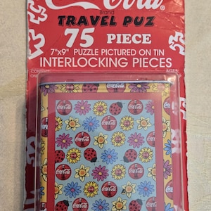 Peut inclure: Un puzzle de voyage de marque Coca-Cola dans un emballage rouge. L'emballage indique "75 pièces" et "Puzzle 7"x9"." La boîte du puzzle présente un motif floral et coccinelle avec des logos Coca-Cola. Le puzzle est composé de pièces imbriquées.