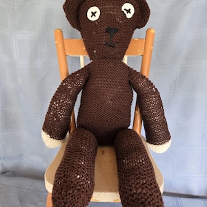 May include: A brown knitted teddy bear with white accents on the paws and button eyes. The bear is seated on a wooden chair. The bear's nose is black, and the eyes are white with black cross stitches. The chair has a light brown finish.