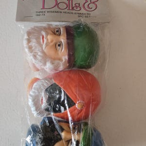Peut inclure: Un paquet de trois têtes de Rois Mages, chacune avec un chapeau de couleur différente : vert, orange et bleu. Les têtes sont en matériau clair et mesurent environ 7 cm de hauteur. L'emballage porte la mention "Dolls &" et "Three Wisemen Heads".