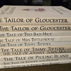 以下が含まれることがあります： 白の表紙に黒い文字で書かれた、子供向けのヴィンテージ本の束。「The Tailor of Gloucester」や「The Tale of Two Bad Mice」などのタイトルが含まれています。本は積み重ねられています。