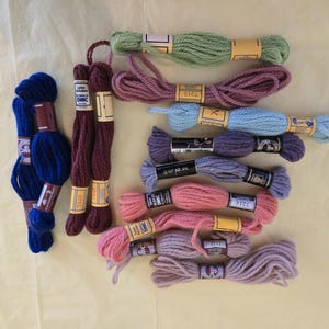 Puede incluir: Una colección de madejas de hilo de bordar de colores en tonos azul, burdeos, verde, morado, rosa y lavanda. El hilo está cuidadosamente dispuesto, con cada madeja etiquetada con una banda de papel. La imagen muestra una variedad de colores y texturas, ideal para proyectos de manualidades.