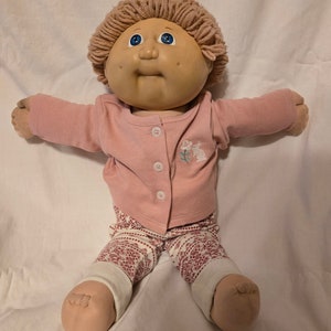 Peut inclure: Une poupée Cabbage Patch Kid avec des cheveux en laine blonde, des yeux bleus et des joues roses. La poupée porte une chemise rose à manches longues avec un motif brodé blanc et un pantalon à motifs rose et blanc.