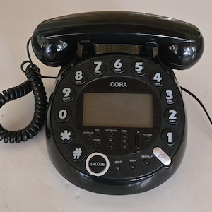 Puede incluir: Teléfono con cable negro con una base redonda y un auricular curvo. El teléfono tiene una pantalla digital y botones para números, funciones y control de volumen. La marca "CORA" es visible.