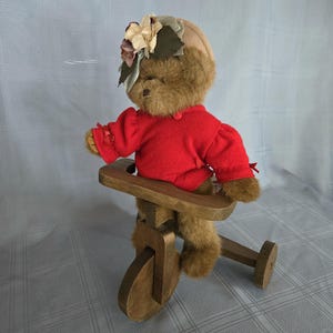 Puede incluir: Un oso de peluche marrón con un suéter rojo y un sombrero floral, montado en un triciclo de madera. El oso está sentado en el triciclo, que tiene una rueda delantera y dos patas traseras.