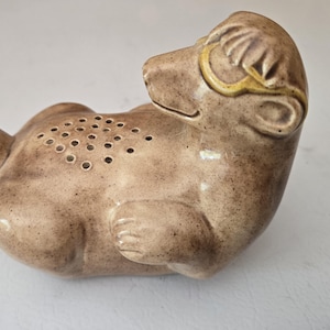 Könnte beinhalten: Keramikfigur eines liegenden Bären in Brauntönen. Der Bär hat eine Reihe kleiner Löcher auf seinem Körper und gelbe Akzente um die Ohren. Es handelt sich wahrscheinlich um einen Salz- oder Pfefferstreuer.