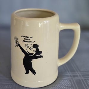 May include: A cream-coloured ceramic beer stein with a side handle. The front features a black graphic of a cartoon bear holding a smaller mug, accompanied by the black text that reads A TOAST TO YOUR SUCCESS! The mug has a smooth, cylindrical body.