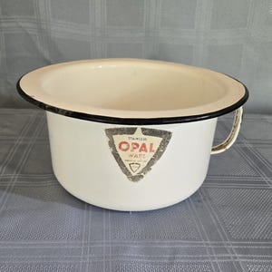 May include: Cream-colored enamelware chamber pot with a black rim and handle. The pot has a triangular sticker with the text "OPAL WARE" and "STAINLESS". The pot is on a blue and gray checkered surface.