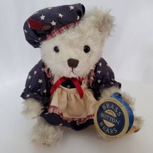 May include: A white teddy bear wearing a blue and white star patterned dress with a red ribbon and a red, white, and blue patterned hat. The bear is sitting on a white surface and has a tag that says "Handcrafted Brass Button Bears Fully Jointed".