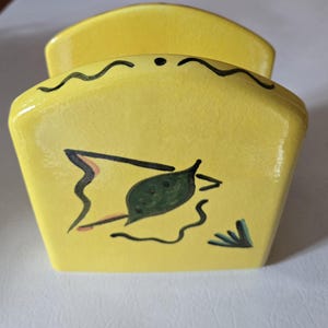 May include: Yellow ceramic napkin holder with a black and green leaf design. The holder has a wavy black line around the top edge.