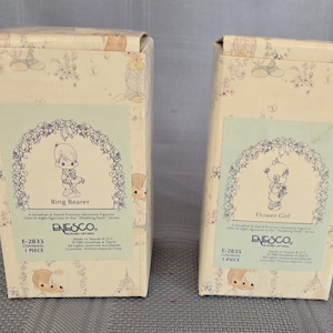 May include: Two decorative boxes with illustrations of children in wedding attire. The boxes are labeled "Ring Bearer" and "Flower Girl" and feature the Enesco logo. The boxes have a light beige background with floral and character designs.