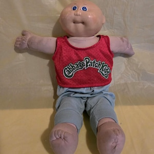 Könnte beinhalten: Cabbage Patch Kids Puppe mit kahlem Kopf, blauen Augen und neutralem Gesichtsausdruck. Die Puppe trägt ein rotes Mesh-Tanktop mit dem Schriftzug "Cabbage Patch Kids" in Grün und eine blaue Jogginghose. Die Arme der Puppe sind ausgestreckt.
