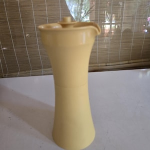 Può includere: Brocca giallo pallido con coperchio e beccuccio. La brocca ha una base svasata e un corpo sottile e curvo. Il coperchio è piccolo e rotondo, e il beccuccio è curvo. La brocca è fatta di un materiale solido.