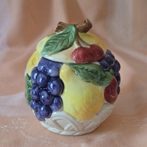 Peut inclure: Un pot en céramique avec un couvercle, décoré de fruits colorés. Le pot présente un motif de raisins, de cerises et de citrons. Le couvercle est surmonté d'un nœud marron et de feuilles vertes. La base du pot a un motif de tissage de panier.