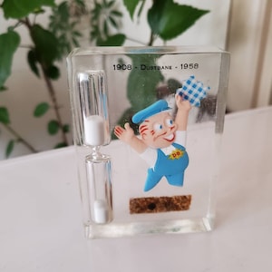 May include: A clear plastic hourglass with white sand inside. The hourglass is in a clear plastic cube with a cartoon character holding a blue and white checked cloth. The text "1908 - Dustbane - 1958" is printed on the cube.