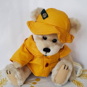 May include: A soft toy teddy bear dressed in a yellow raincoat and hat. The bear has cream-coloured fur, black eyes, and a black nose. The hat features a small green patch and a silver button. The raincoat is fastened with a red button.