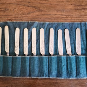 May include: A collection of twelve silver butter knives, each with a rounded blade and floral engraving, presented in a teal fabric roll. The knives are displayed on a wooden surface, possibly indicating a vintage or antique set.