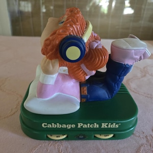May include: A Cabbage Patch Kids figurine wearing headphones, dressed in pink, white, and blue. The figurine is on a green base with the words "Cabbage Patch Kids" and two dials. The figurine is in a listening pose.