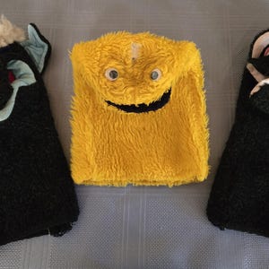 May include: Three plush hand puppets. Two are black with white and light blue accents, and one is yellow with a smiling face. The puppets have a shaggy texture and are designed to fit over a hand.