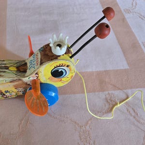 May include: Vintage wooden pull toy of a bee with a yellow and brown body, black antennae, and orange wings. The bee has a large, round eye and a crown on its head. The toy is labelled "Queen Busy Bee".