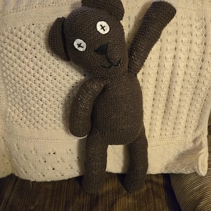 May include: A handmade brown knitted teddy bear with white accents on the eyes and arm. The bear has a black stitched nose and mouth. The bear is resting on a cream-colored, patterned blanket.