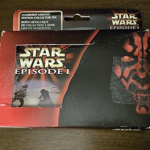 May include: A Star Wars Episode I collector's tin. The box is red and black, featuring the Star Wars logo and imagery of Darth Maul. The packaging includes text in English and French, indicating a limited edition.