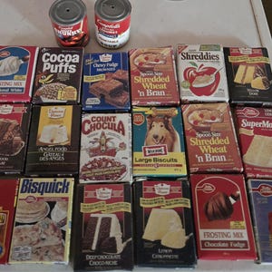 Può includere: Una collezione di scatole e lattine di cibo vintage, tra cui preparati per torte, preparati per brownies e cereali per la colazione. I marchi includono Duncan Hines, Betty Crocker e Campbell's. Le scatole presentano grafica e testo colorati.