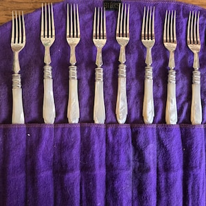 May include: A collection of antique forks with silver tines and decorative handles, presented in a deep purple fabric roll. The handles have a pearlescent finish, and the tines are neatly arranged. These forks are a vintage dining accessory.