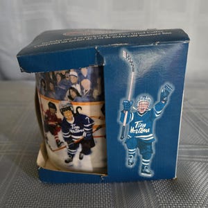 May include: A dark blue cardboard box containing a ceramic mug featuring a hockey-themed graphic. The box displays a child in a blue Tim Hortons hockey jersey holding a stick, with text stating it includes one 57g packet of fine grind coffee.