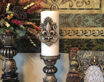 May include: Two ornate candle holders with a white candle in one. The candle holder has a decorative fleur-de-lis design. The candle holders are gold and black with a distressed finish.
