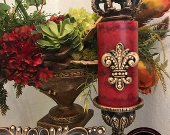 May include: A red candle with a gold crown and fleur-de-lis design on top, sitting on a gold candle holder. The candle is surrounded by a floral arrangement with red, green, and orange flowers. The candle holder has a gold base with a decorative design. The candle is labeled "Your Majesty Decor".