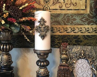 May include: Two ornate gold and black candle holders with a white pillar candle in one. The candle holder has a decorative gold design on the top. The background is a patterned fabric with gold and brown colors.