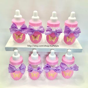 Puede incluir: Ocho biberones de pl&aacute;stico transparente con tapas rosas y tetinas blancas. Cada botella tiene una etiqueta rosa festoneada con un gr&aacute;fico de mariposa dorada y un lazo de sat&eacute;n lavanda. El texto http://etsy.com/shop/AJPetals es visible.