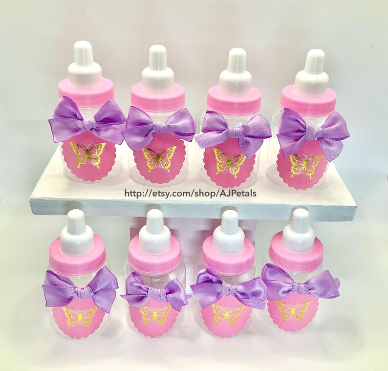 Puede incluir: Ocho biberones de pl&aacute;stico transparente con tapas rosas y tetinas blancas. Cada botella tiene una etiqueta rosa festoneada con un gr&aacute;fico de mariposa dorada y un lazo de sat&eacute;n lavanda. El texto http://etsy.com/shop/AJPetals es visible.